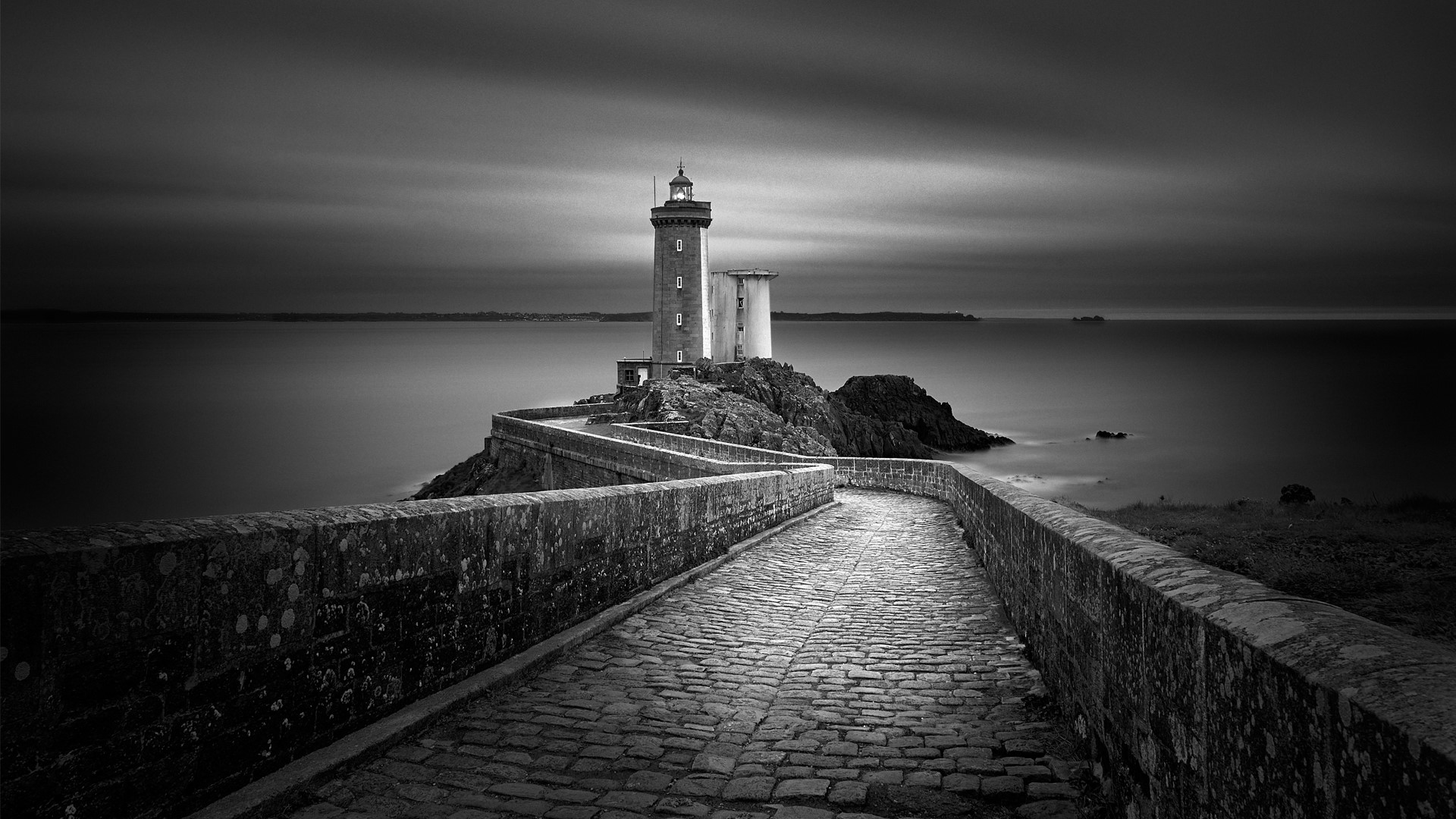 Footpath leading towards lighthouse at sea shore against cloudy sky at dusk, Brittany, France